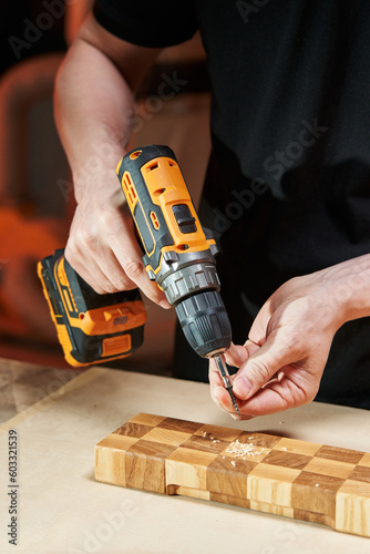 The hands of a Caucasian worker install a self-tapping screw into the bit of an electric screwdriver with a replaceable battery to screw it into a wooden part