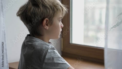 Teenager stays next to window alone and is thinking about something at home. Sad boy is looking out of the window. Emotional concept