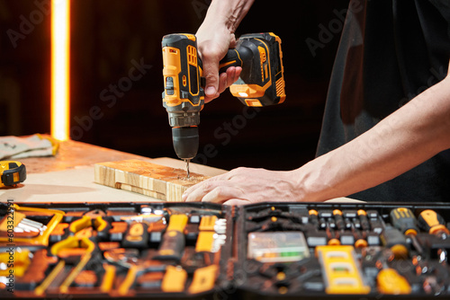 The hands of a Caucasian worker drill with an electric drill-driver with a replaceable battery a wooden part on a workbench, on which lies a case with different tools