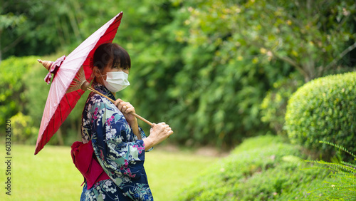 Cute Japanese women wearing beautiful traditional clothes dress walking relax happily in casual Yukata kimonos with white masks and red umbrellas in a fresh green natural garden.