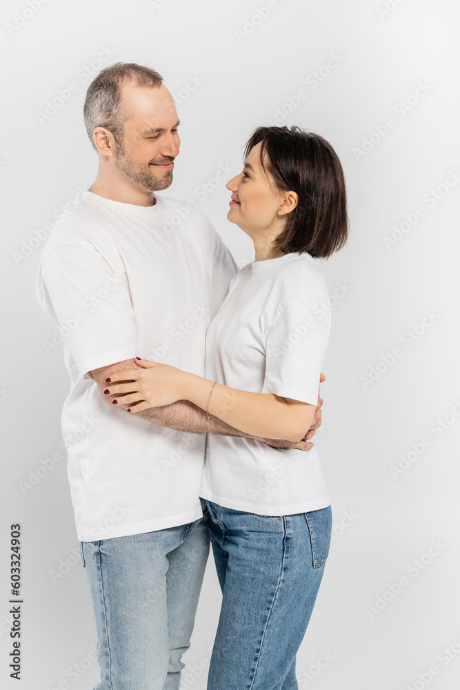 cheerful and tattooed woman with short brunette hair hugging joyous husband with bristle while standing together in white t-shirts and looking at each other isolated on grey background, happy couple cheerful and tattooed woman with short brunette hair hugging joyous husband with bristle while standing together in white t-shirts and looking at each other isolated on grey background, happy couple