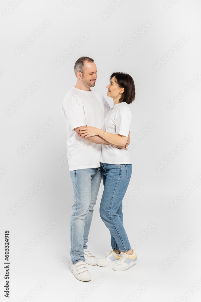 full length of smiling woman with short brunette hair hugging husband with bristle while standing together in white t-shirts and denim jeans looking at each other on grey background, happy couple full length of smiling woman with short brunette hair hugging husband with bristle while standing together in white t-shirts and denim jeans looking at each other on grey background, happy couple