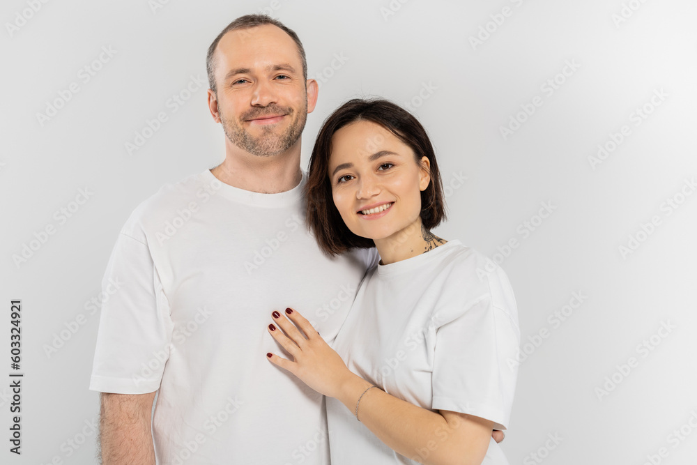 cheerful and tattooed woman with short brunette hair hugging joyous husband with bristle while standing together in white t-shirts and looking at camera isolated on grey background, happy couple cheerful and tattooed woman with short brunette hair hugging joyous husband with bristle while standing together in white t-shirts and looking at camera isolated on grey background, happy couple