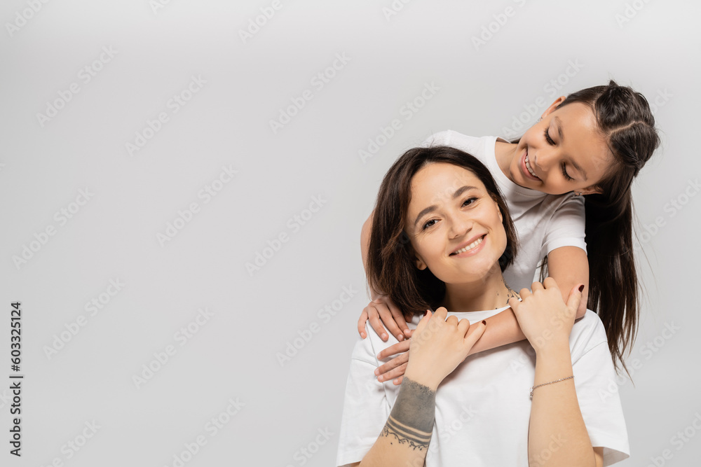 happy preteen daughter with brunette hair embracing cheerful mother with tattoo on hand and looking at camera on grey background, Child protection day, mother and daughter happy preteen daughter with brunette hair embracing cheerful mother with tattoo on hand and looking at camera on grey background, Child protection day, mother and daughter