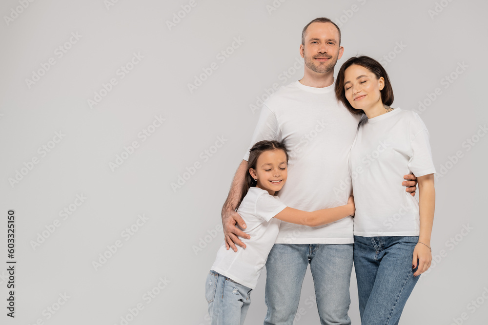 happy preteen girl hugging father near mother with tattoo while standing together in blue denim jeans and white t-shirts and looking at camera on grey background, Happy children's day happy preteen girl hugging father near mother with tattoo while standing together in blue denim jeans and white t-shirts and looking at camera on grey background, Happy children's day