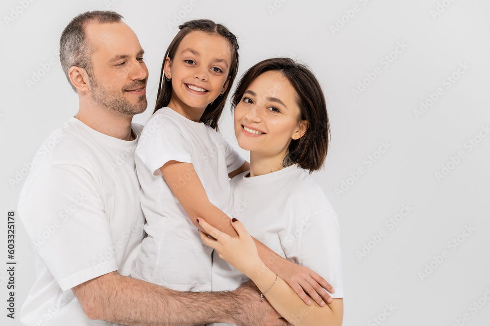portrait of happy family in white t-shirts looking at camera and hugging each other on grey background, International child protection day, parents and daughter portrait of happy family in white t-shirts looking at camera and hugging each other on grey background, International child protection day, parents and daughter