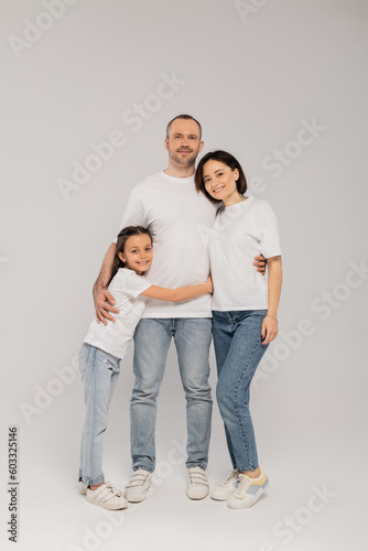 joyous preteen girl hugging father near mother with tattoo while standing together in blue denim jeans and white t-shirts and looking at camera on grey background, Happy children's day,