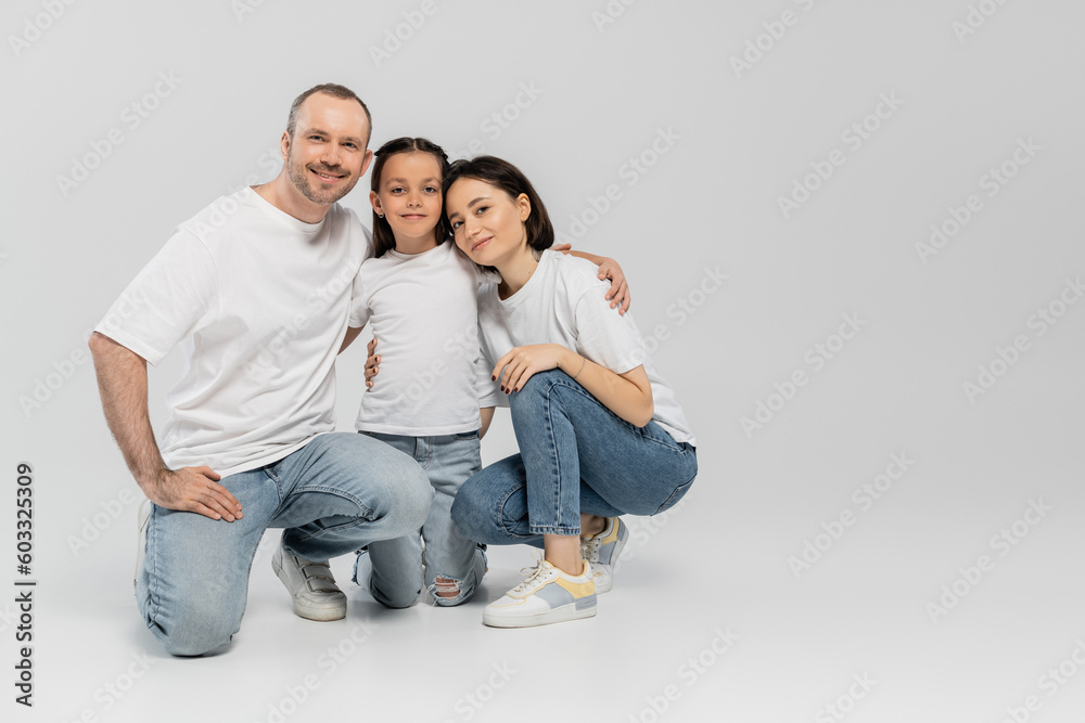 happy father and mother with short hair hugging cheerful preteen daughter while sitting together in white t-shirts and blue denim jeans on grey background, Happy children's day, June 1 happy father and mother with short hair hugging cheerful preteen daughter while sitting together in white t-shirts and blue denim jeans on grey background, Happy children's day, June 1