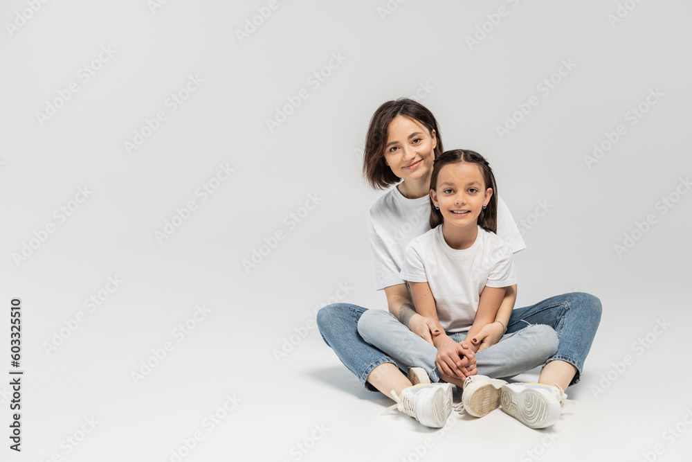 positive mother with tattoo and short hair hugging preteen brunette daughter while sitting together in white t-shirts and blue denim jeans on grey background, child protection day