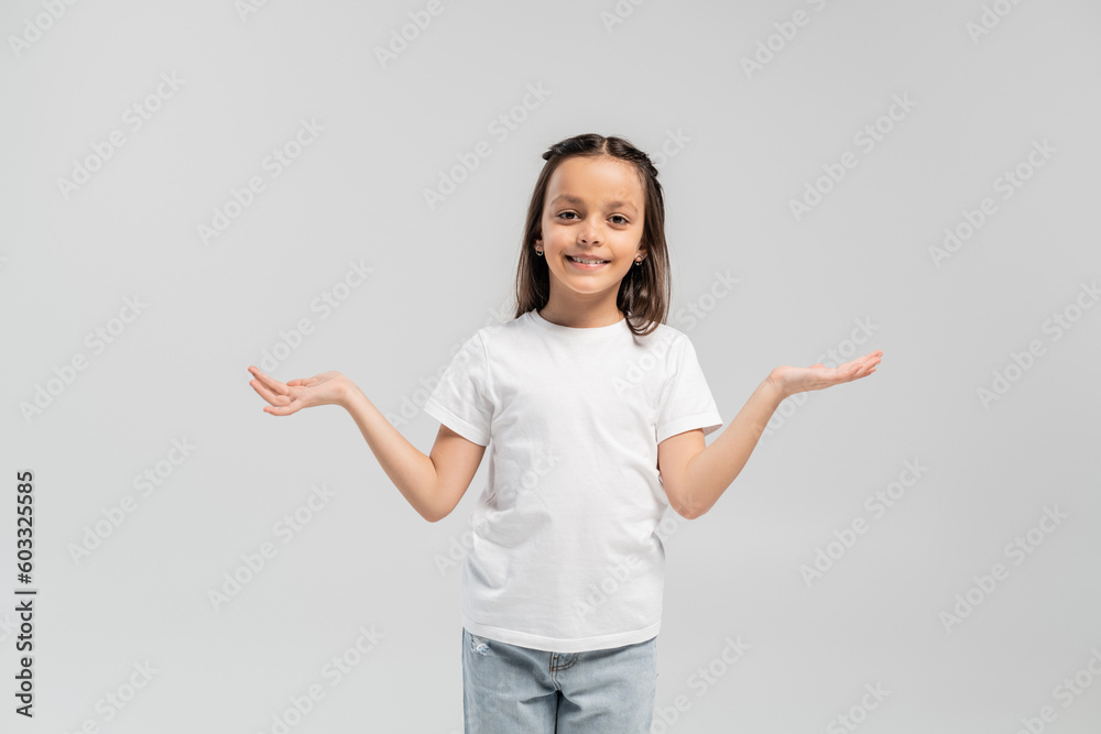Smiling brunette preteen kid in white t-shirt and jeans looking at camera and showing shrug gesture while celebrating child protection day and standing isolated on grey Smiling brunette preteen kid in white t-shirt and jeans looking at camera and showing shrug gesture while celebrating child protection day and standing isolated on grey