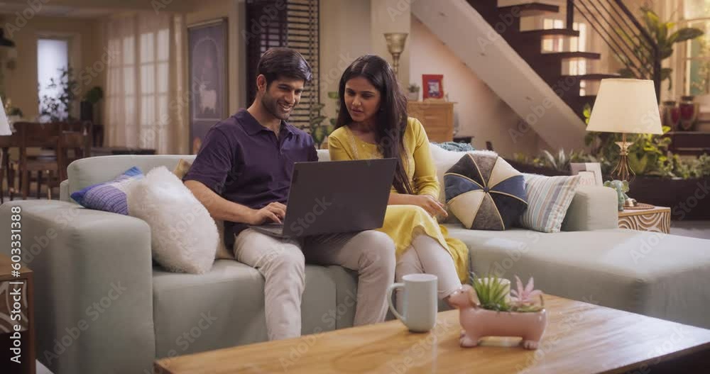 Portrait of Loving Young Indian Couple Enjoys Browsing Online Shopping Websites Together On A Laptop, While Sitting Cozily On Their Living Room Sofa. Browsing Interner, Choosing Products to Purchase