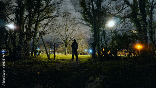 A threatening mysterious hooded figure. Silhouetted by street lights on the edge of town. Standing on the edge of wasteland. On a spooky winters night.