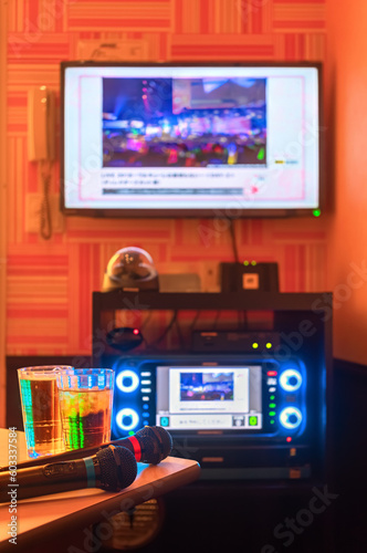 Two black microphones for singing songs on a corner of a table next to two drink glasses in a Japanese karaoke club room with a controller lit by backlit LEDs on buttons under a TV screen on the wall.