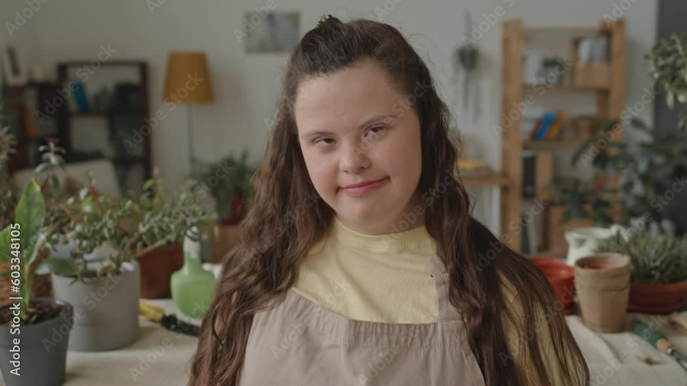 Tilt up shot of girl with Down syndrome wearing apron standing in floristic studio in front houseplants on table, holding digital tablet and posing for camera with smile Tilt up shot of girl with Down syndrome wearing apron standing in floristic studio in front houseplants on table, holding digital tablet and posing for camera with smile