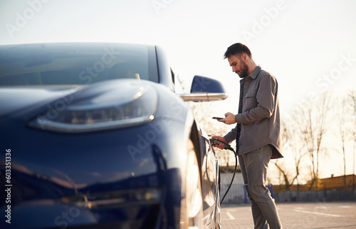 View from the side. Charging the automobile. Man is standing near his electric car outdoors
