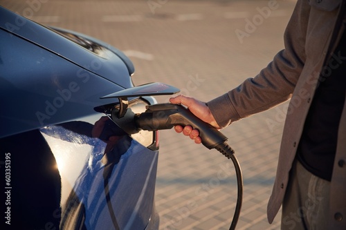 Blue colored automobile. Putting the charger in. Man is standing near his electric car outdoors