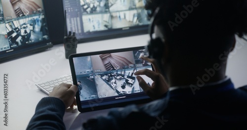 Male operator monitors CCTV cameras. African American man uses digital tablet with view of security cameras. Computer screens on background. Tracking and monitoring system. Concept of social safety.