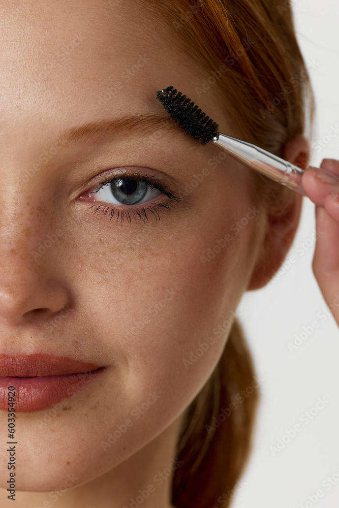 Close-up portrait of beautiful young redhead female model with freckles, brushing brows against white studio background. Concept of natural beauty, cosmetology, cosmetics, skin care