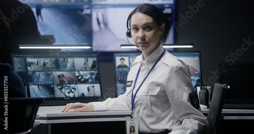 Female operator in headset in police monitoring center turns and looks at camera. Computer monitors and big digital screen with surveillance CCTV cameras video footage on background. Tracking system.