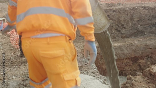 UK - cement pours into a pre-dug pit. Two workers supervise. When the cement dries, it will form the base of a house foundation.