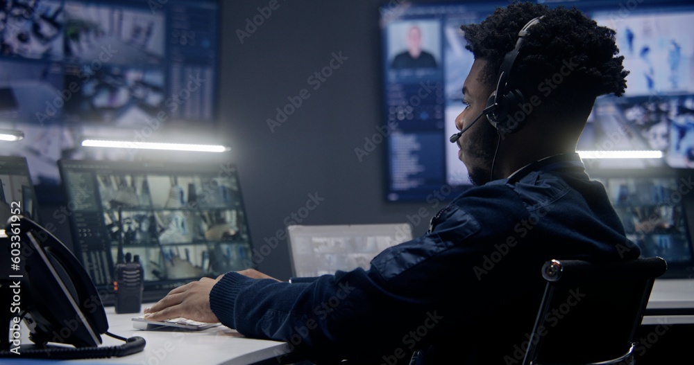 African American male operator in headset monitors security cameras with AI face scanning in police surveillance room. Multiple big screens with CCTV footage at background. Concept of social safety.