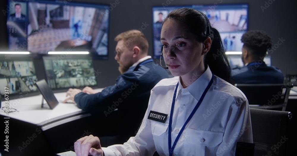 Obraz premium Close up shot of female security operator in headset looking at monitors with displayed CCTV cameras in police surveillance center. Multi ethnic colleagues work at background. Social safety concept.