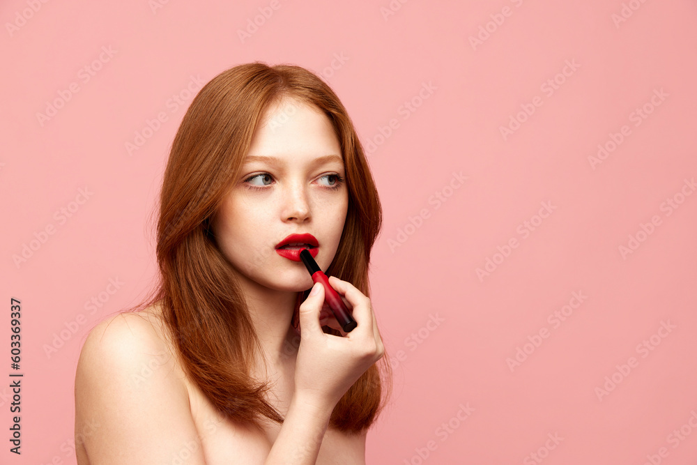 Portrait of young, beautiful, redhead girl with well-kept skin, applying red lipstick against pink studio background. Concept of natural beauty, makeup, cosmetology, cosmetics, skin care Portrait of young, beautiful, redhead girl with well-kept skin, applying red lipstick against pink studio background. Concept of natural beauty, makeup, cosmetology, cosmetics, skin care