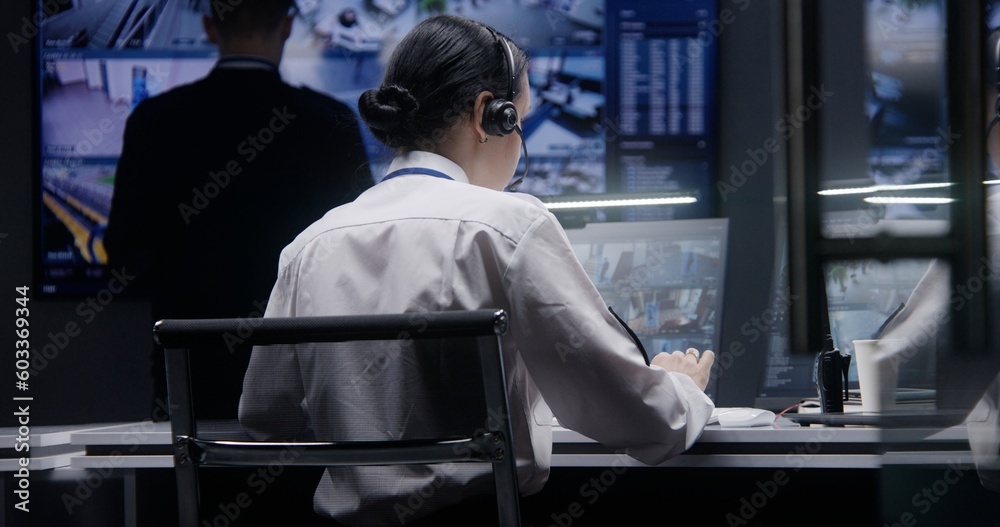 Female security officer works on computer in police monitoring center, controls CCTV cameras with AI facial recognition. Male worker looks at big digital screen with surveillance camera video footage.