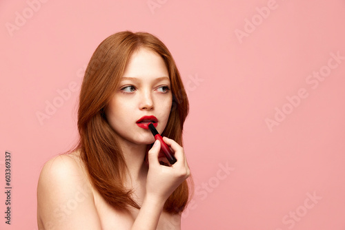 Portrait of young, beautiful, redhead girl with well-kept skin, applying red lipstick against pink studio background. Concept of natural beauty, makeup, cosmetology, cosmetics, skin care