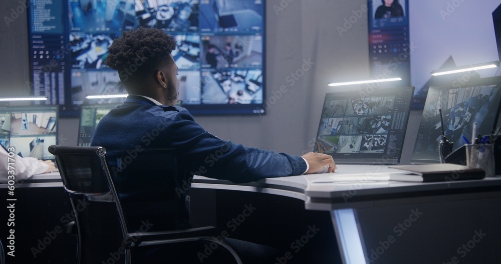 African American security operator monitors CCTV cameras with AI face recognition. Multiple big screens on the wall showing security cameras view. Colleague works at background. Social safety concept. African American security operator monitors CCTV cameras with AI face recognition. Multiple big screens on the wall showing security cameras view. Colleague works at background. Social safety concept.