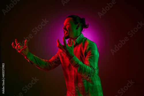 Portrait of emotional man in checkered shirt shouting in anger and irritation against dark gradient studio background in neon light. Concept of human emotions, facial expression, lifestyle