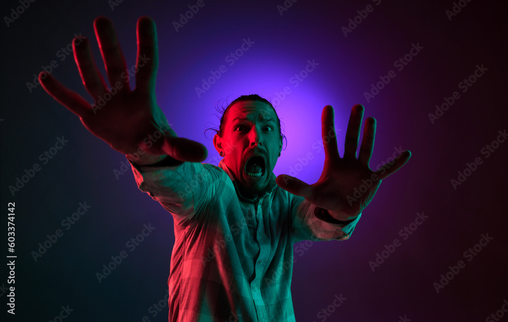 Portrait of emotional young man showing fearful face against dark gradient studio background in neon light. Horror. Concept of human emotions, facial expression, lifestyle Portrait of emotional young man showing fearful face against dark gradient studio background in neon light. Horror. Concept of human emotions, facial expression, lifestyle