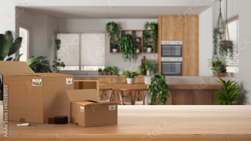 Wooden table, desk or shelf with stack of cardboard boxes over blurred view of kitchen and dining room, houseplants interior design, moving house concept with copy space © ArchiVIZ