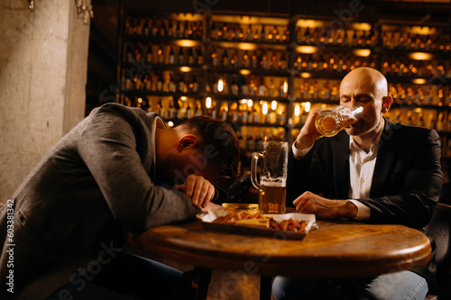 A young man in a suit sleeps near a glass of whiskey and beer on a table in a pub, another man drinks alcohol