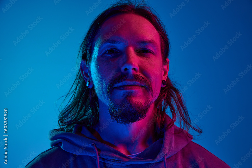 Portrait of man with moustache in casual clothes, hoodie looking at camera, posing with smile against blue studio background in neon light. Concept of human emotions, facial expression, lifestyle - obrazy, fototapety, plakaty 