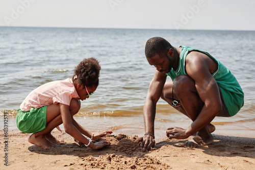 African American dad building sand castle together with his little daughter while they spending time on the beach