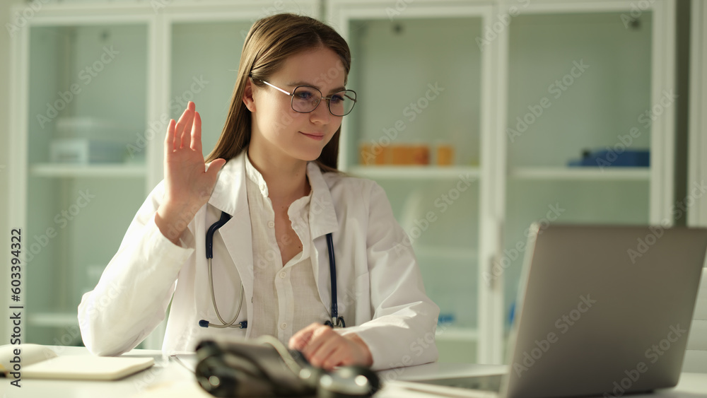 Woman doctor in glasses waving hand at laptop screen in clinic. Telemedicine concept Woman doctor in glasses waving hand at laptop screen in clinic. Telemedicine concept