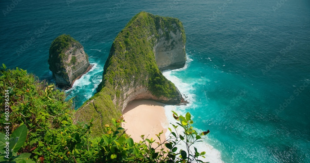 Blue ocean waves crashing on tropical rocky cliffs and sandy Kelingking beach. Top down view of turquoise sea water, green vegetation in Bali.