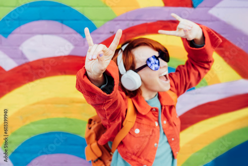 Focus on rock and roll hand gesture of emotional stylish woman wearing wireless headphones, listening to music and singing on rainbow brick wall background. Music festival. hipster lifestyle.