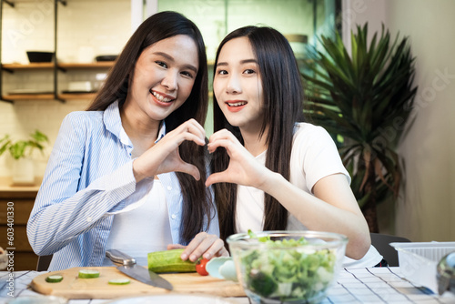 Young Asian couple LGBTQ woman smiling and making heart sign gesture together while cooking salad and leaning cooking on table at home.