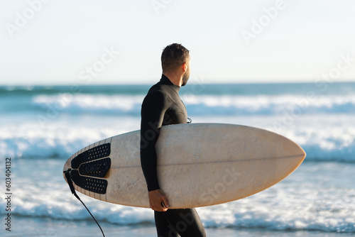 A man surfer in a wetsuit walking on the shore holding a surfboard and looking at the sea