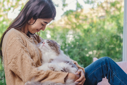 Fototapeta Woman with long hair in brown sweater and jean holding persain cat in her lap and embrassed him with love feelling, girl and cat looking each other with love and care emotion