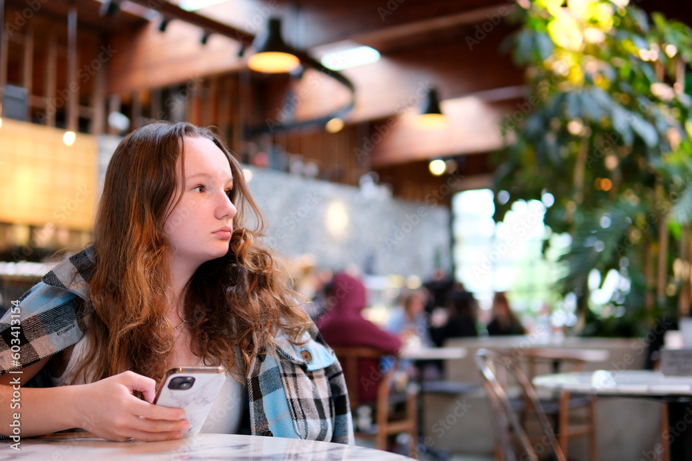Young woman hold the phone in your hands throw a checkered blue jacket over your shoulders in city cafe.Drink coffee, wait for somebody. High quality photo