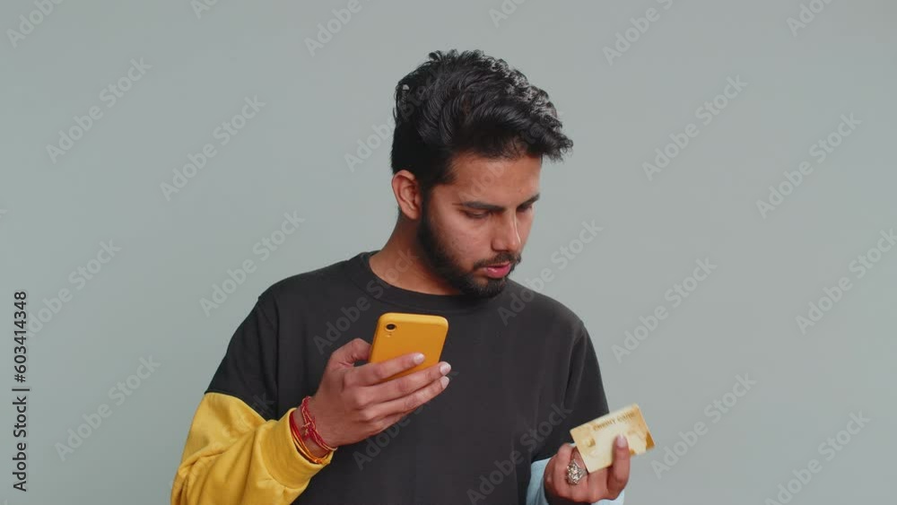 Sincere indian man customer using credit bank card and smartphone while transferring money, purchases online shopping, payment. Finance and internet. Handsome guy isolated alone on gray background Sincere indian man customer using credit bank card and smartphone while transferring money, purchases online shopping, payment. Finance and internet. Handsome guy isolated alone on gray background