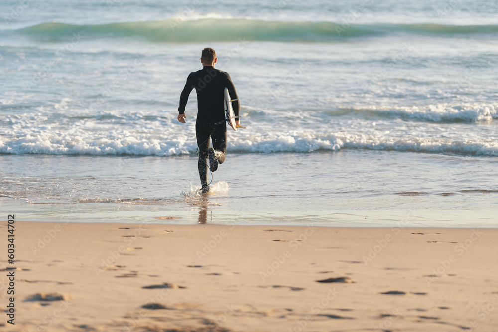 An athletic man surfer in a wetsuit running to the ocean holding a surfing board An athletic man surfer in a wetsuit running to the ocean holding a surfing board