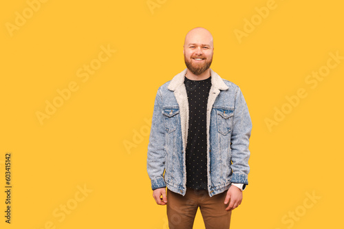 Studio portrait of a wide smiling bald bearded man wearing jeans jacket over yellow background.