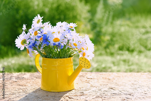 meadow flowers bouquet in yellow watering can on table, natural sunny background. rustic floral composition in garden. summer blossom season. copy space. template for design. photo