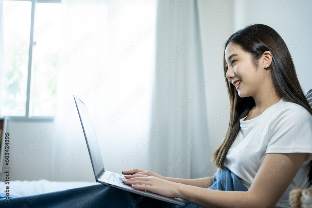 Young smiling Asian woman in casual pajamas outfit sitting in bed in morning working on laptop, freelancer at home. Young smiling Asian woman in casual pajamas outfit sitting in bed in morning working on laptop, freelancer at home.