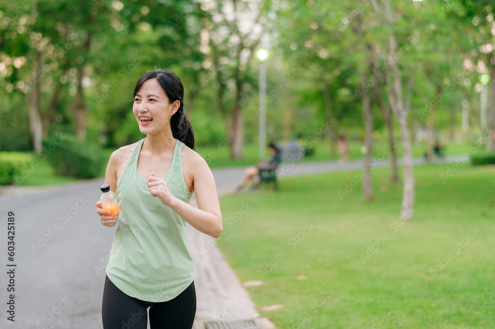 Female jogger. Fit young Asian woman with green sportswear drinking organic orange juice after running and enjoying a healthy outdoor. Fitness runner girl in public park. Wellness being concept Female jogger. Fit young Asian woman with green sportswear drinking organic orange juice after running and enjoying a healthy outdoor. Fitness runner girl in public park. Wellness being concept