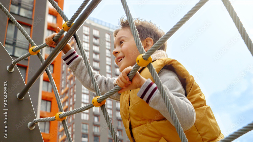 Cute smiling boy climbing up the rope net on the public playground at park. Active child, sports and development, kids playing outdoors Cute smiling boy climbing up the rope net on the public playground at park. Active child, sports and development, kids playing outdoors